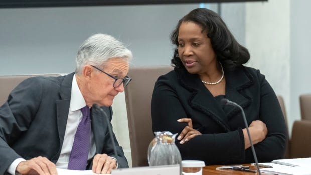 A dark-complected woman and a grey-haired Caucasian man are shown in conversation while seated at a table.