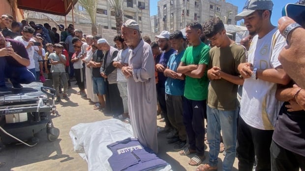 A row of people stand by a white shroud on the ground with a press vest on top of it. The people have their arms folded and their heads bowed, and appear to be in the middle of speaking or reciting a prayer together. 
