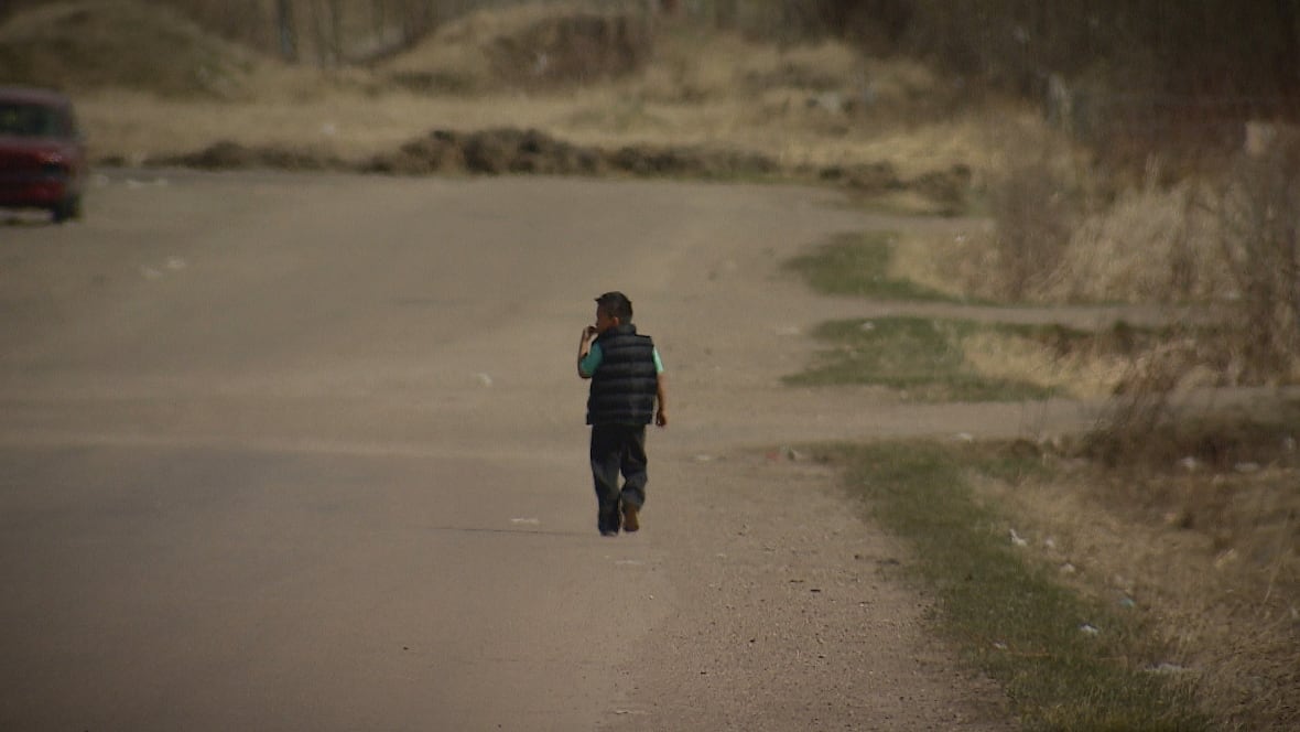 A young boy in a black vest and green shirt walks down a dirt road away from the camera.