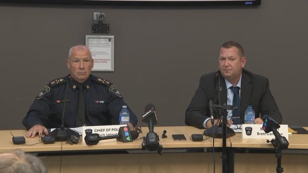 A uniformed, middle-aged police chief and a police sergeant in a black suit sit at a table in a conference room with microphones in front of them, listening to a reporter's question
