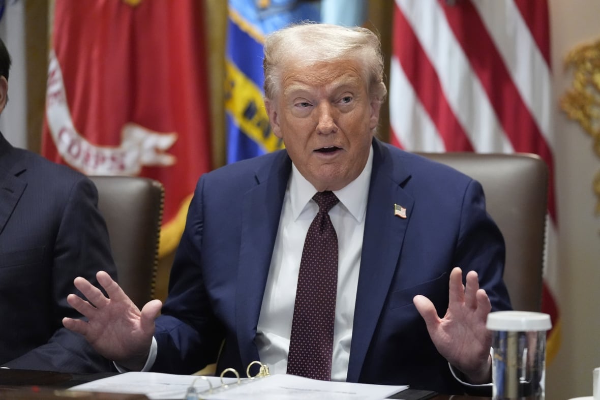 The elderly man wears a suit and tie with his hands while speaking. Sitting on a table.