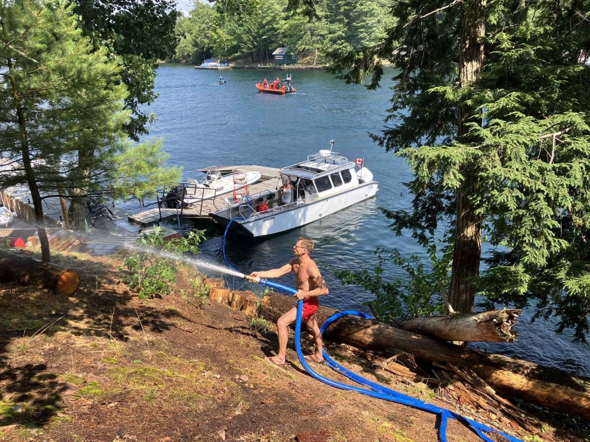 A man wearing flip flops and red swim shorts sprays a hose which leads to a pump on a dock, next to a boat.