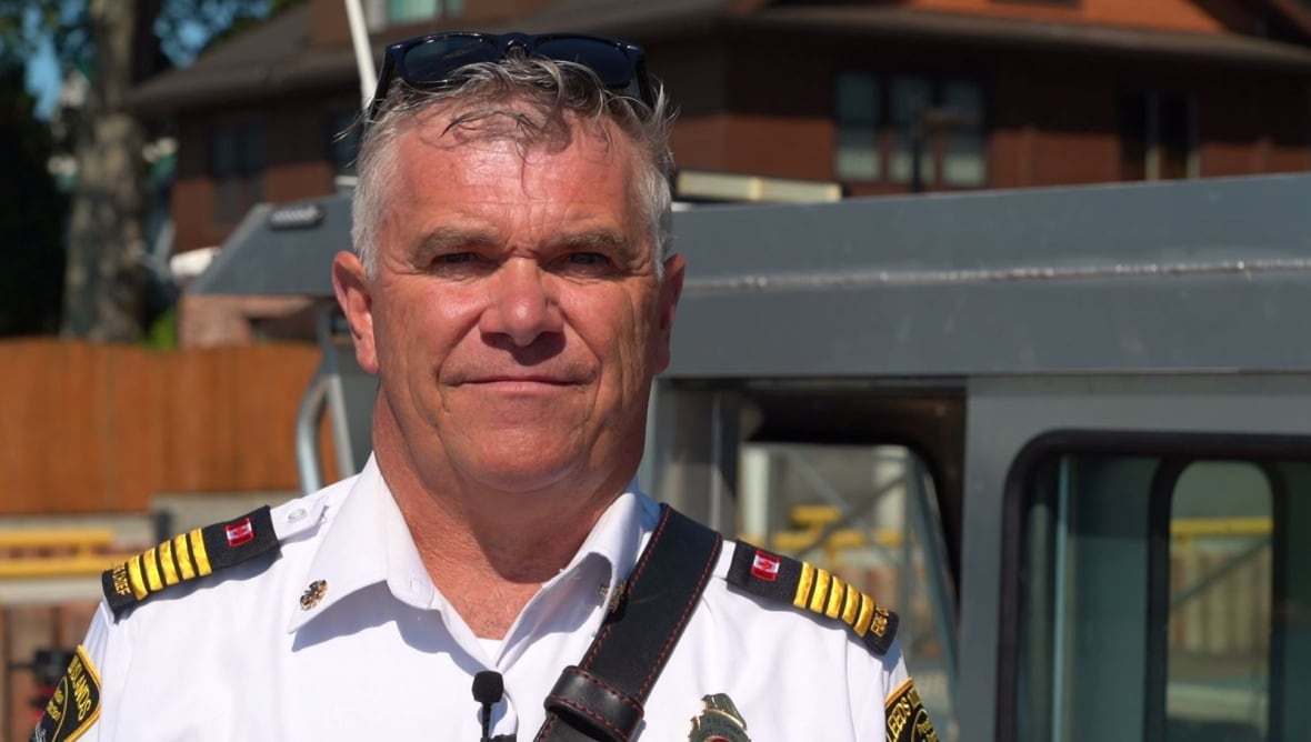 A man in a white uniform shirt, with epaulettes and a badge stands on a dock on a sunny day.