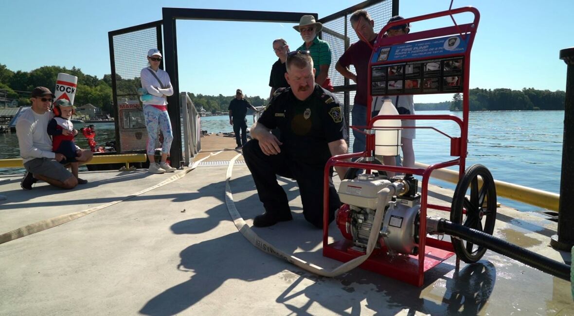 A man in a firefighter uniform works with a red and white pump while and hose, while a crowd of cottagers looks on.