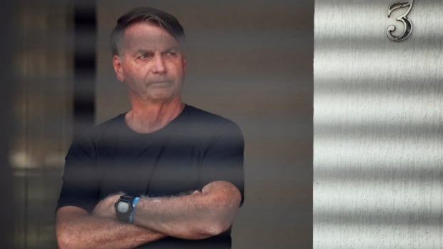 A man in a dark tee-shirt stands looking out a window, with arms crossed in front of his chest.