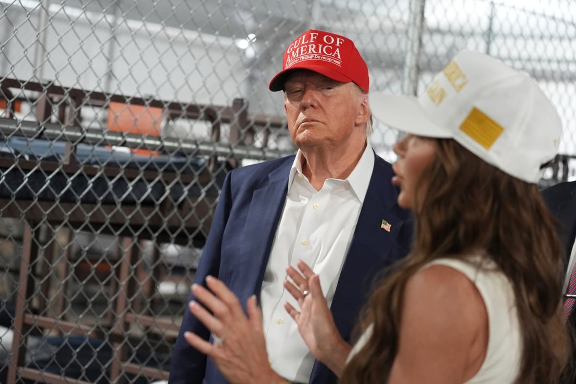 A man and a woman in baseball caps speak to each other inside a facility. He wears a suit jacket and a white shirt, and she is wearing a sleeveless summit.