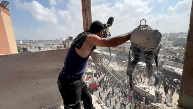 A man on an upper floor of a bombed out building holds up camera gear to show it to people gathered in the square below the building.