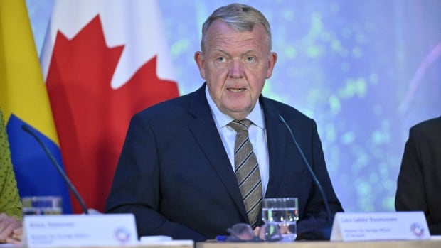 An older cleanshaven man is shown seated and speaking in front of flags, while wearing a suit and tie. 