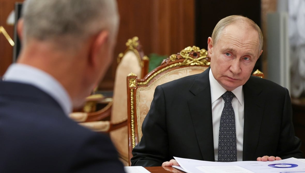 A man wearing a suit sits on a table, looking forward to a person in the unclear introduction, whose head is only visible on the left side of the image.