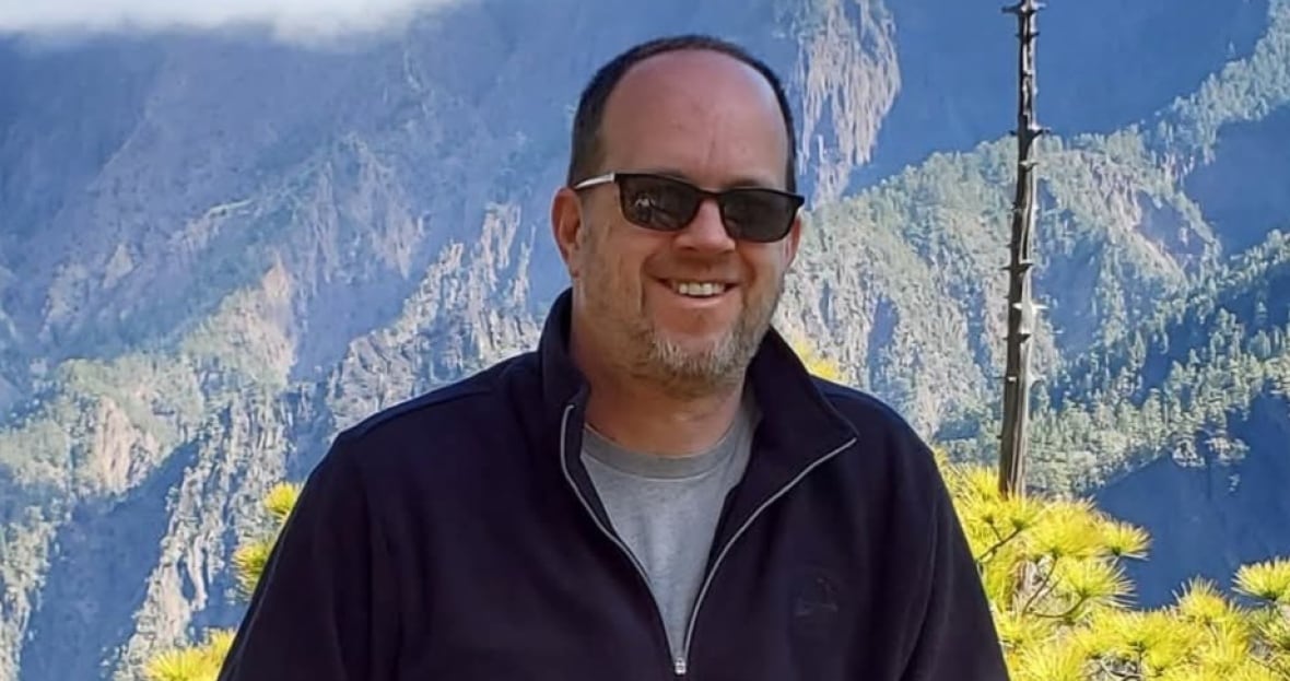 A man stands leans on a wooden railing with mountains and foliage visible behind him