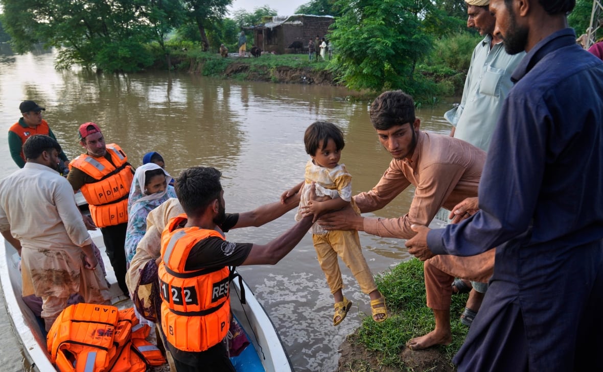 Two men hand over a young child, one to the other. One of the men in a small boat with many other men in orange jackets. The other man who holds the child stands on a herb, with the presence of many other men around him. The water is visible behind them.
