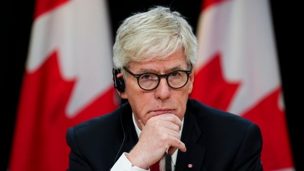 A man in a suit and tie holds his hand to his chin as he sits in front of a microphone. Canadian flags are drapped in the background.