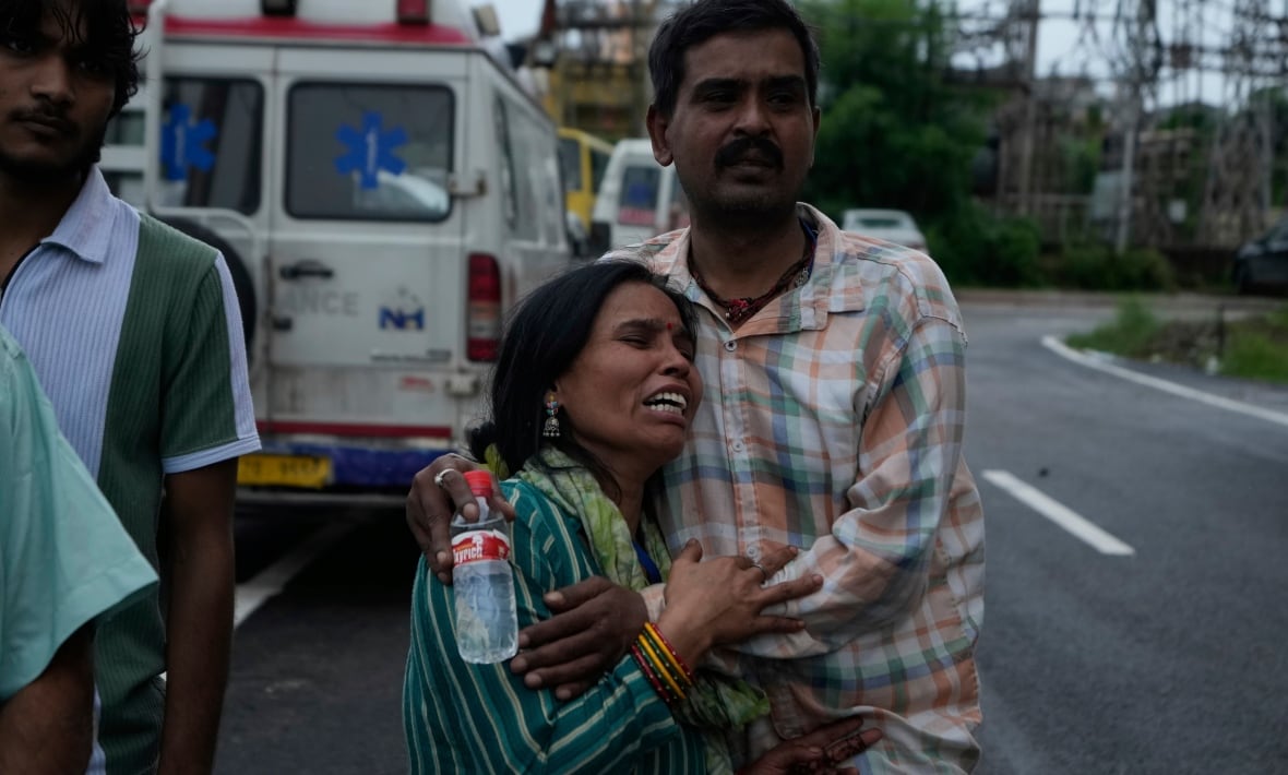 A woman crying as a man holds. They stand on a way with the visible vehicles behind them.