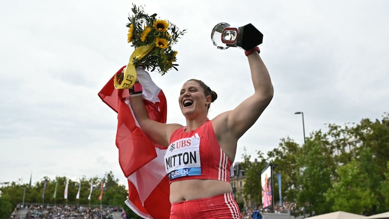 A women's shot putter smiles and holds a trophy and flowers in the air in celebration as she poses for a photograph.