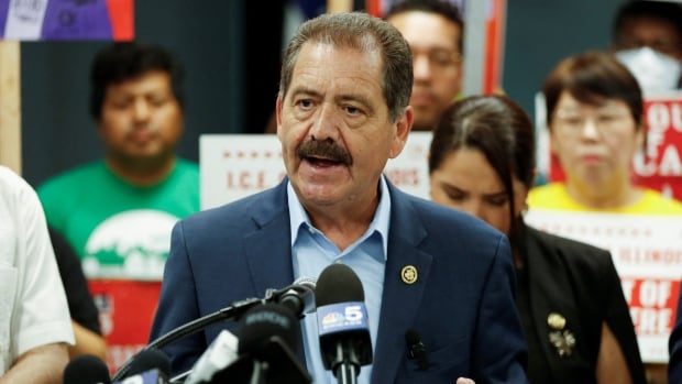 An adult man wearing a blue suit speaks in front of news microphones, with people holding signs seen in the background.