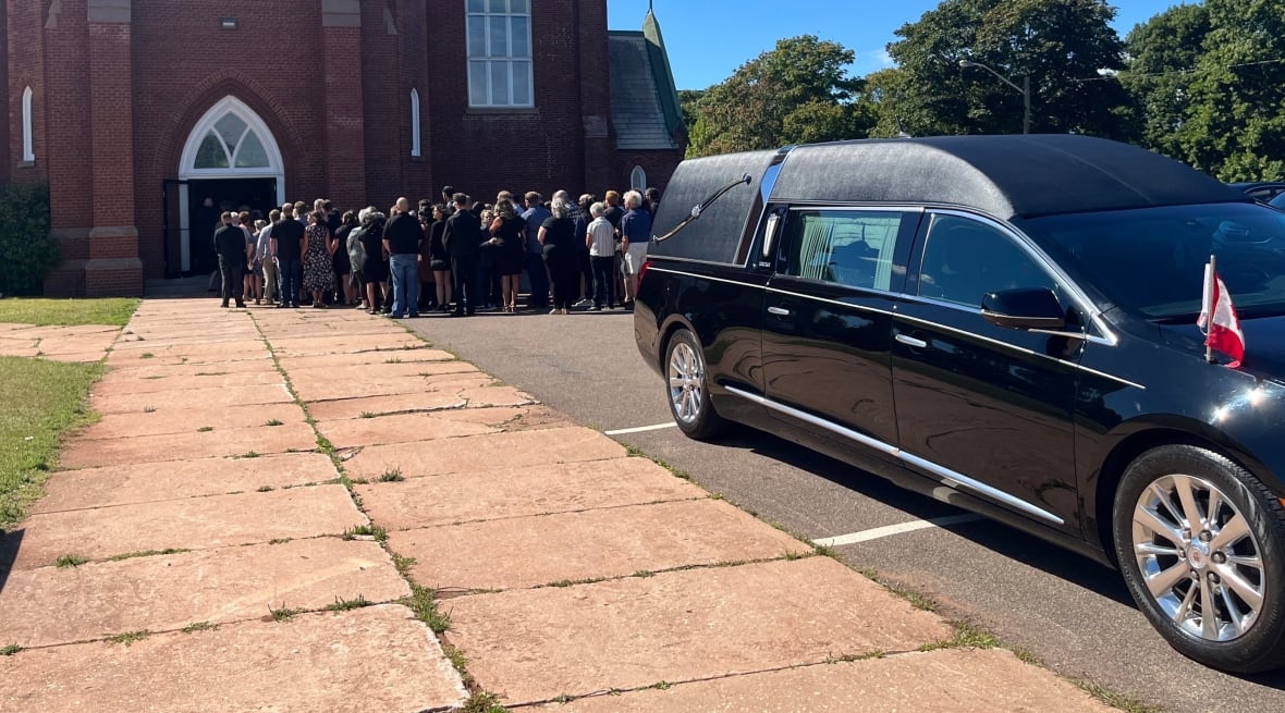 A hearse is parked outside a church and a long line of people waits to get inside.