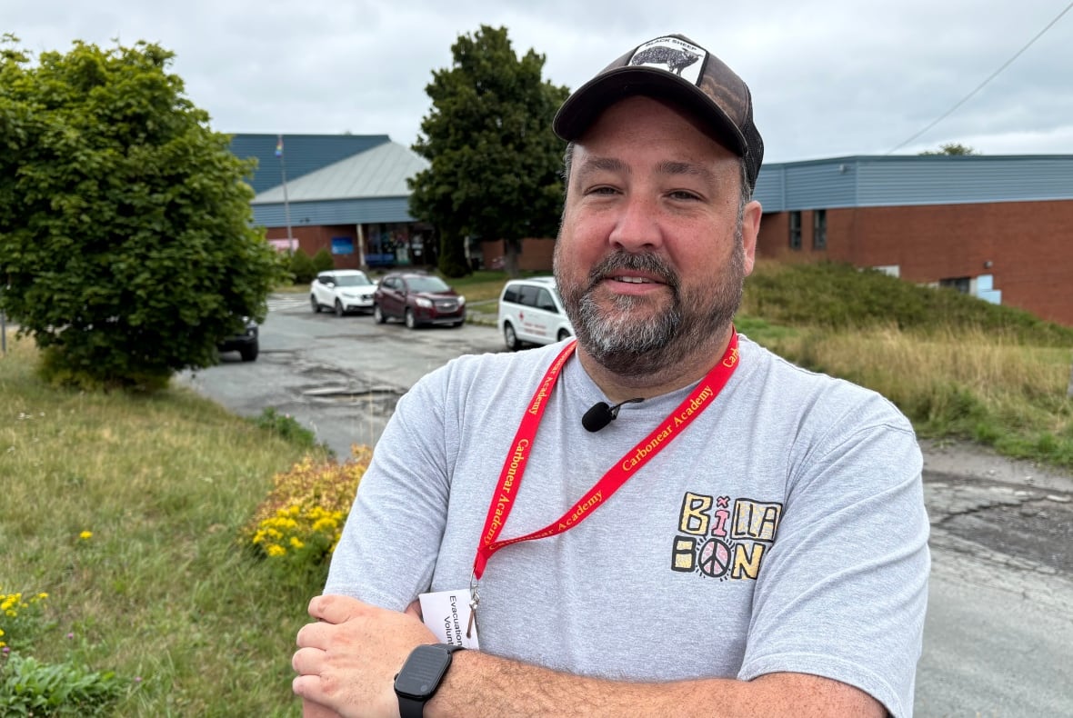 A bearded man in a t-shift and ballcap stands outside, with tall grassy hills, a low building and cars seen on the road behind him.