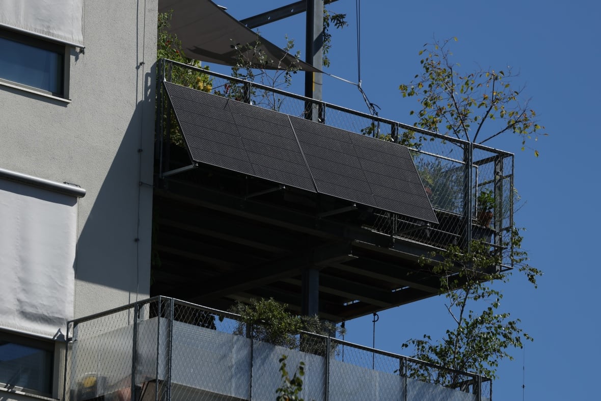 A solar panel hangs from the side of a balcony on a top floor of a gray concrete apartment building.