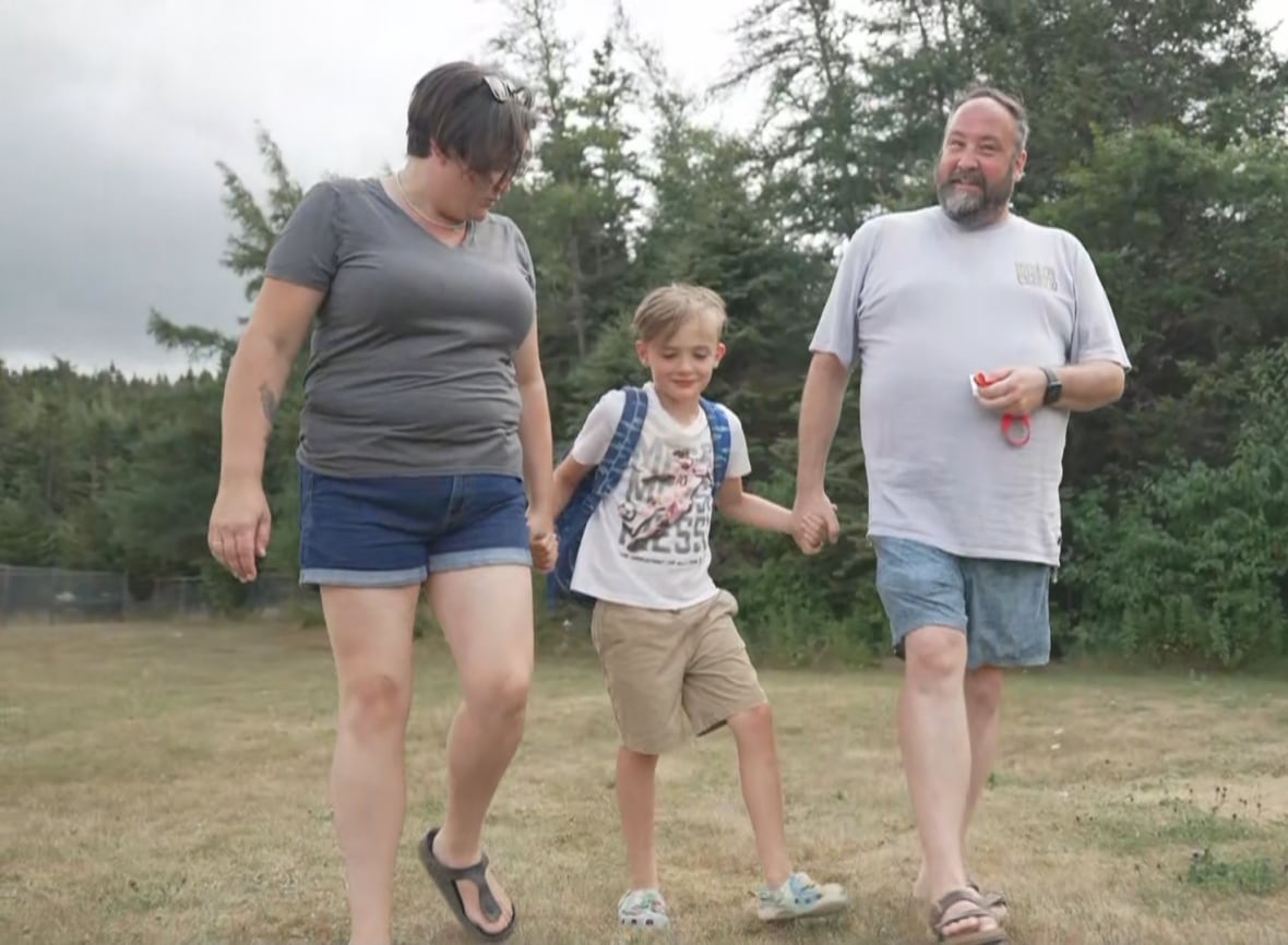 Una donna e un uomo, entrambi vestiti casualmente con magliette e pantaloncini, camminano mano nella mano in un campo, con un ragazzo in mezzo a loro. Dietro il trio compaiono degli alberi sempreverdi.