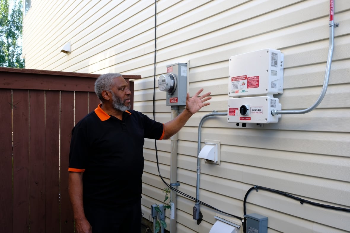 A man stands beside electricity meters and panels mounted on the side of his home.