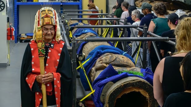 A chief leads a group of people walking around a totem pole laying down, waiting to be transported. 