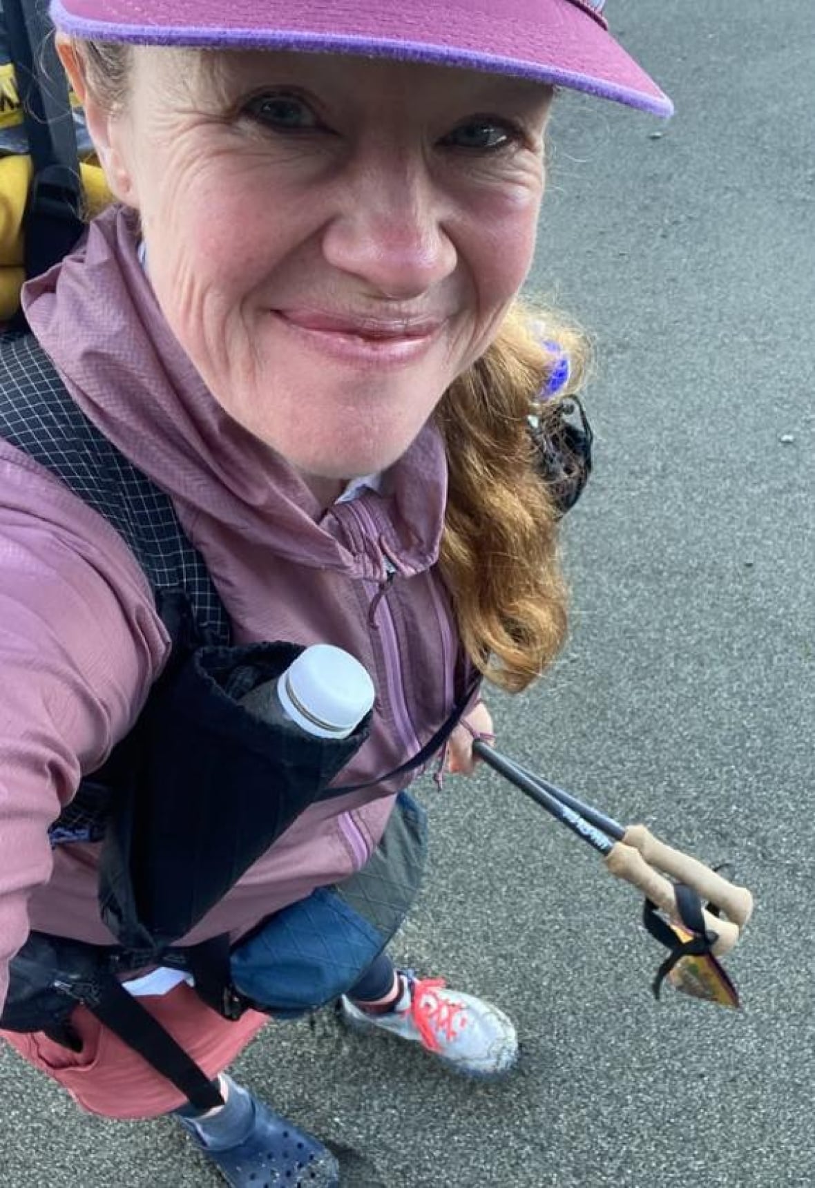 A woman wearing purple hiking gear, including a running shoe and a croc, smiles in a selfie.
