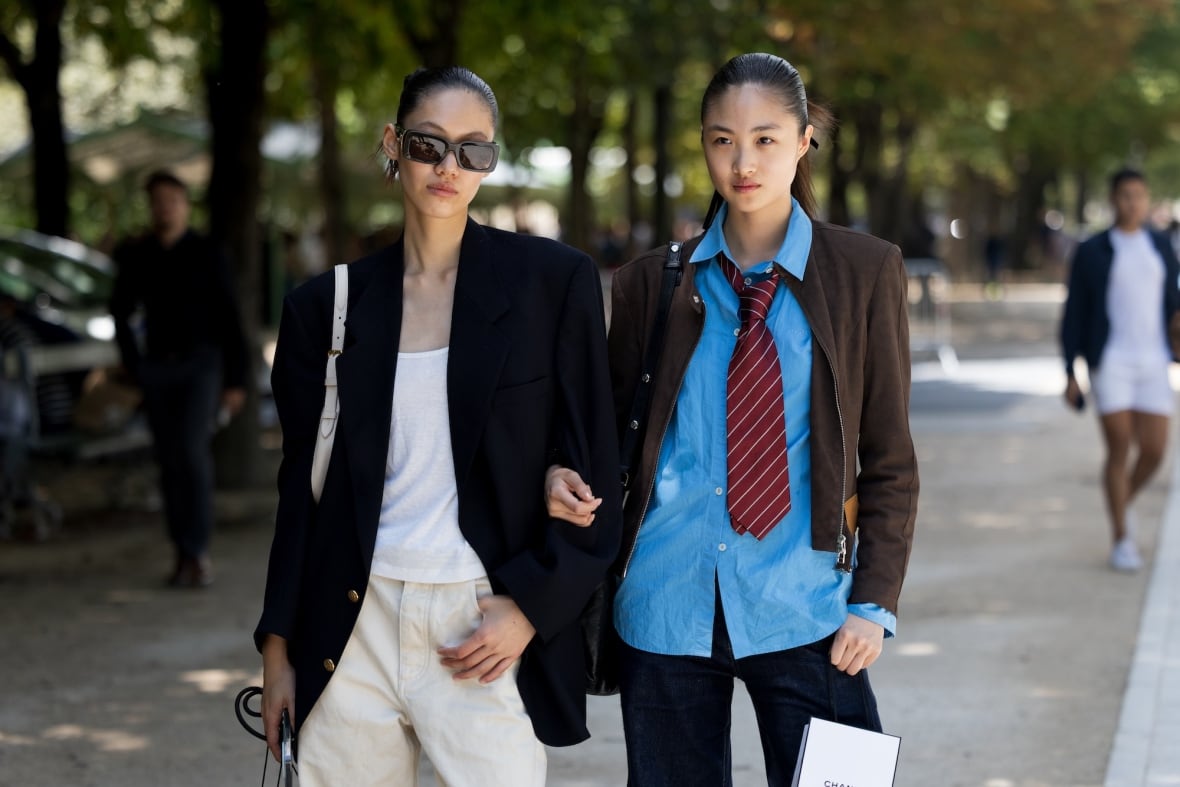 Deux femmes marchant à Paris. À gauche, porte un blazer noir, un débardeur blanc et un pantalon. À droite se trouve une veste en daim marron, une chemise bleue, une cravate à rayures rouges et un jean.