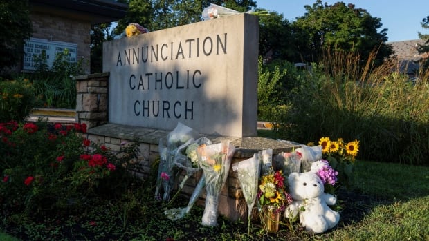 Flowers and teddy bears are seen propped up against a stone sign that reads Anunciation Catholic Church