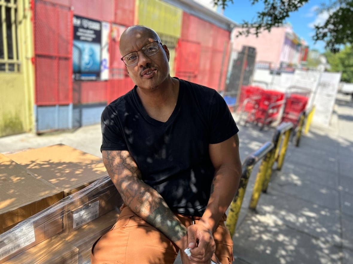 Damon is an American man of African origin with a bald head. He wears rust -colored goods and a back. He sits on a metal iron next to the box cage. Behind it there is a grocery store with red and yellow straps in the front.
