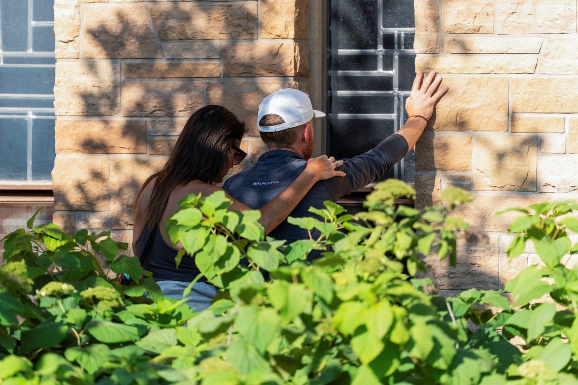 Two people stand in the face of the church window, one with her arm around the shoulder of the other, one with his arm on the wall. 