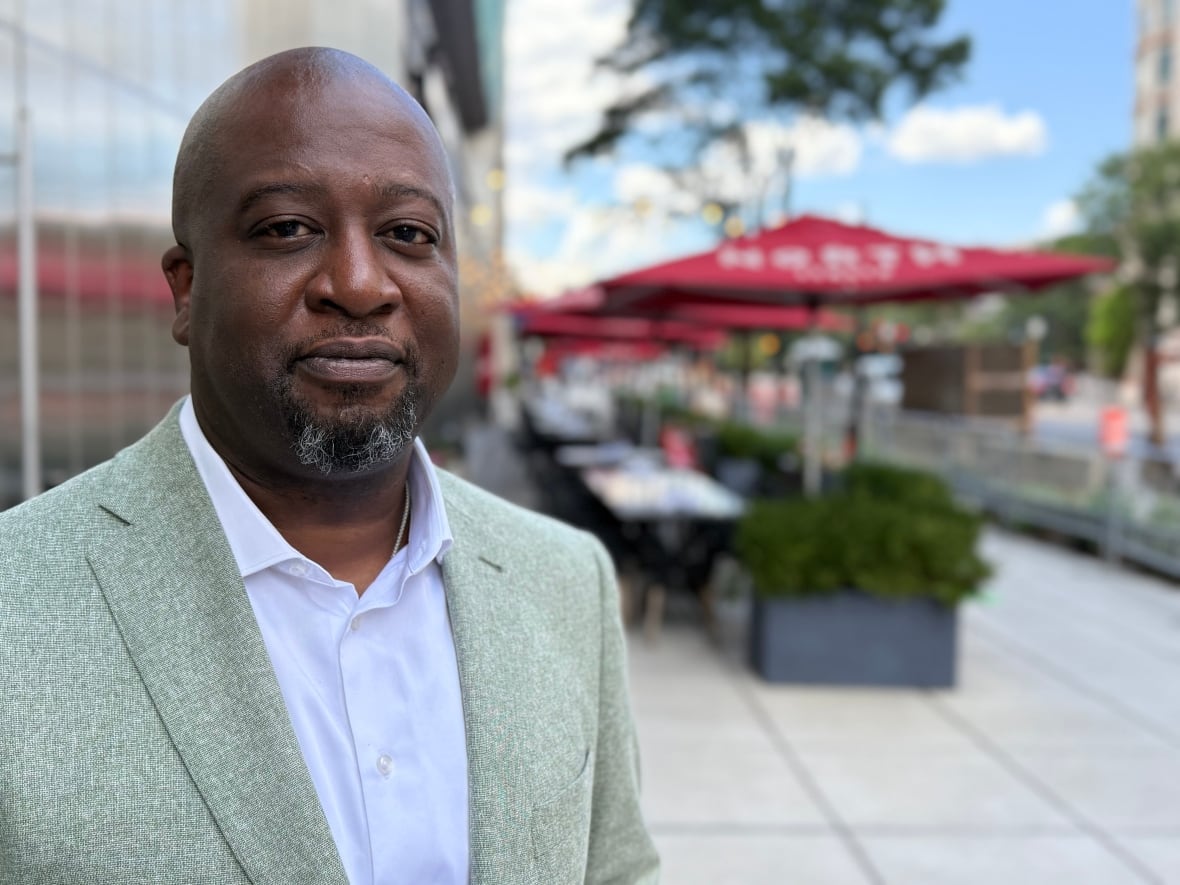 Shawn is a balanced African American man and a dark and gray gaatee. He wears a bright gray suit with a white shamped shirt with the first open button. He stands in front of empty patio with red bugs on the tables.