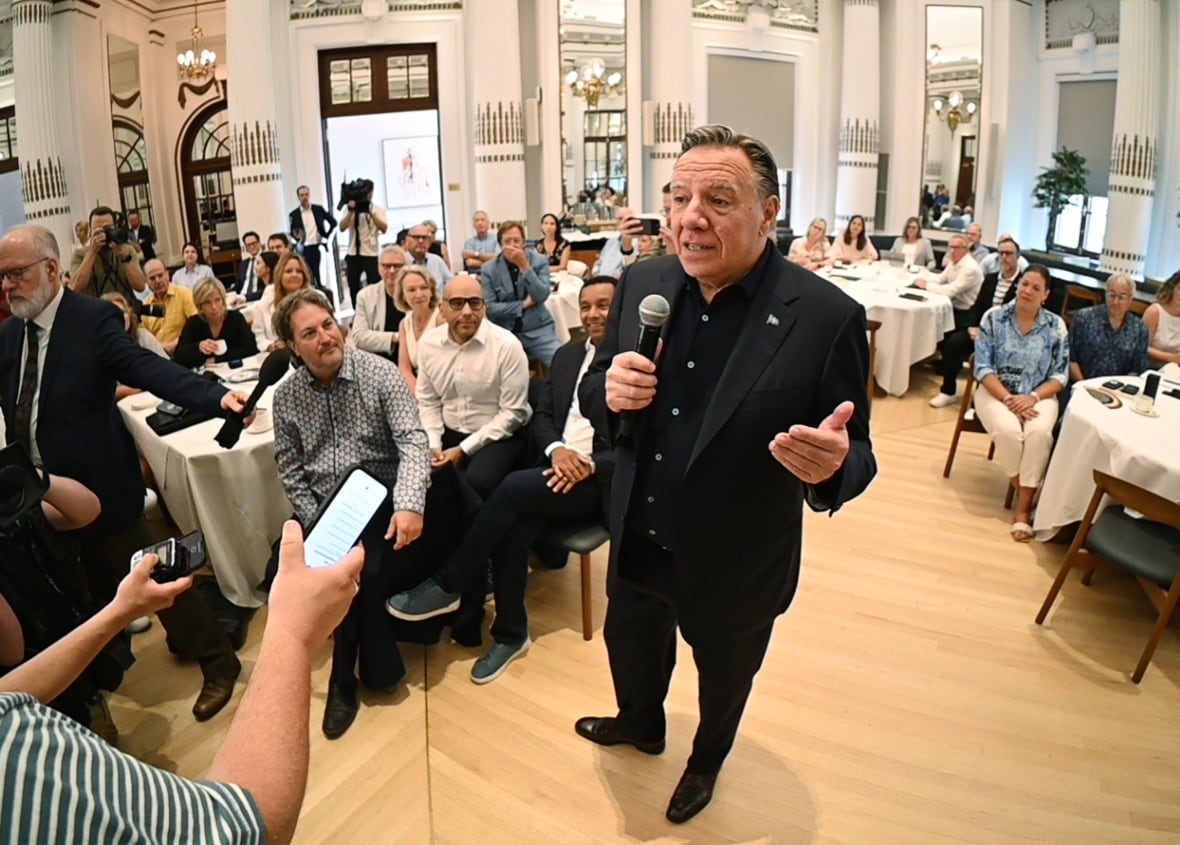 Legault speaking into a microphone amid a crown of people sitting at tables. 