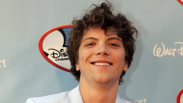 a young man in a blue suit crosses his arms and smiles at the camera on a red carpet