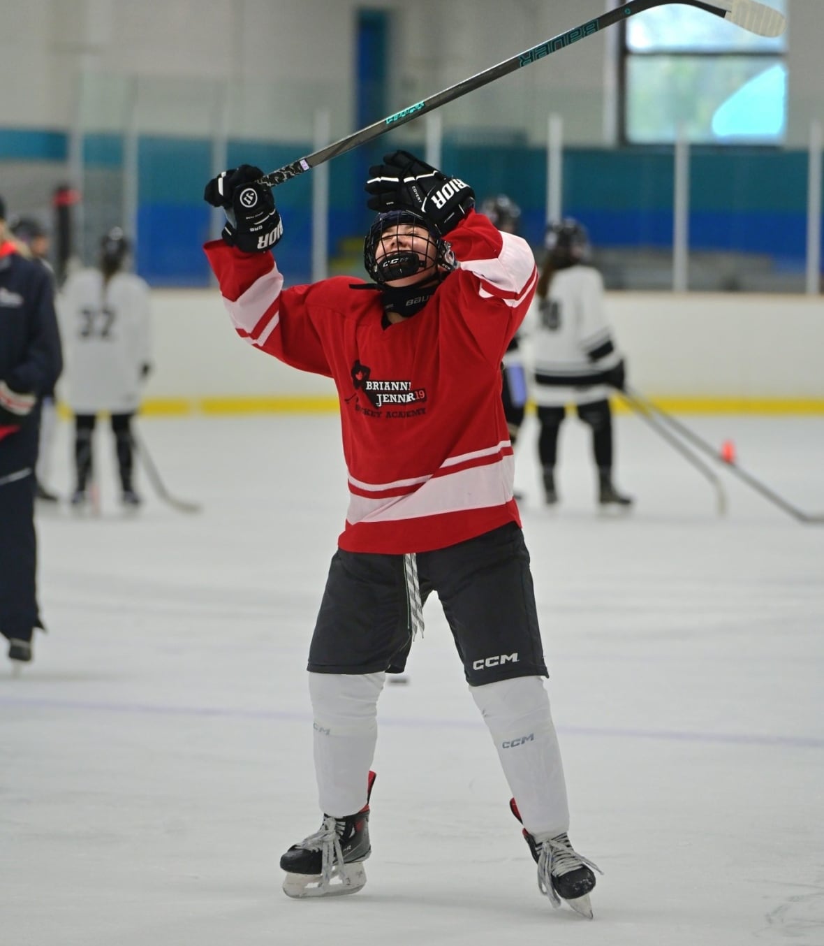 A girl on the ice in hockey equipment and a red jersey, and hands and stick thrown above her head in what appears to be celebration.