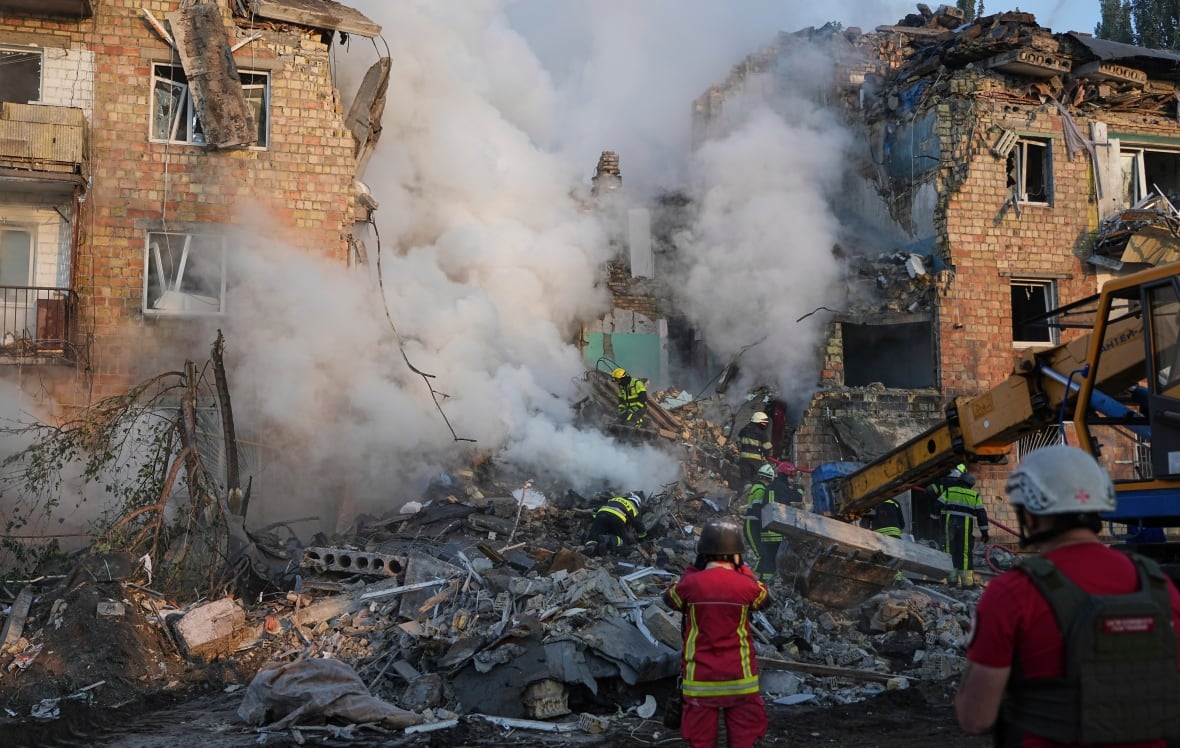Firefighters can be visible at the bottom of the image, with smoke or spray escalating around the central opening in a row of damaged buildings. 