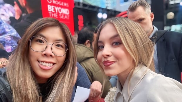 A young woman in glasses poses for a selfie with actress Sydney Sweeney on a busy red carpet in downtown Toronto during daytime in September