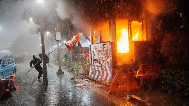 Protestors with flags run by a building that's on fire, in the rain