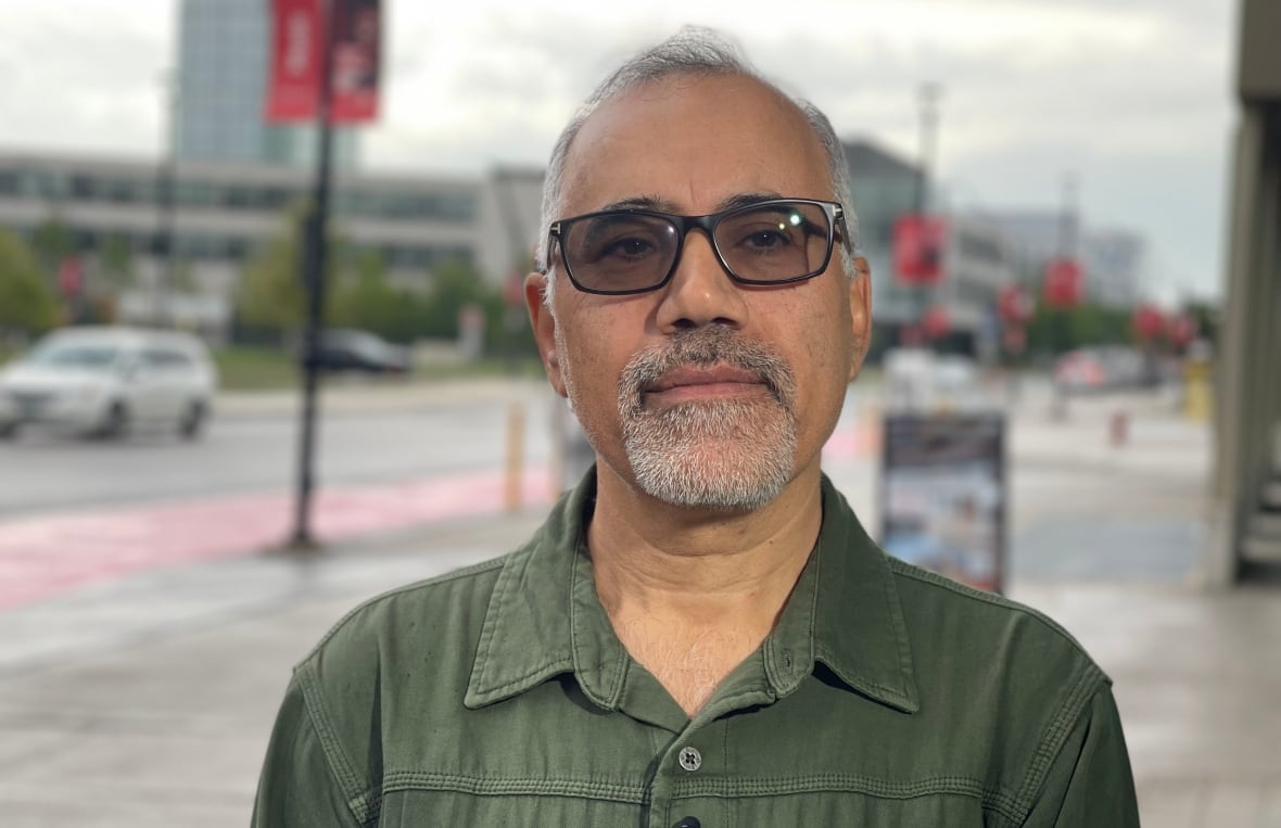 A man in glasses and an olive shirt stands outdoors on an overcast day, with a car driving on a quiet road, red signs on tall posts and buildings seen behind him.