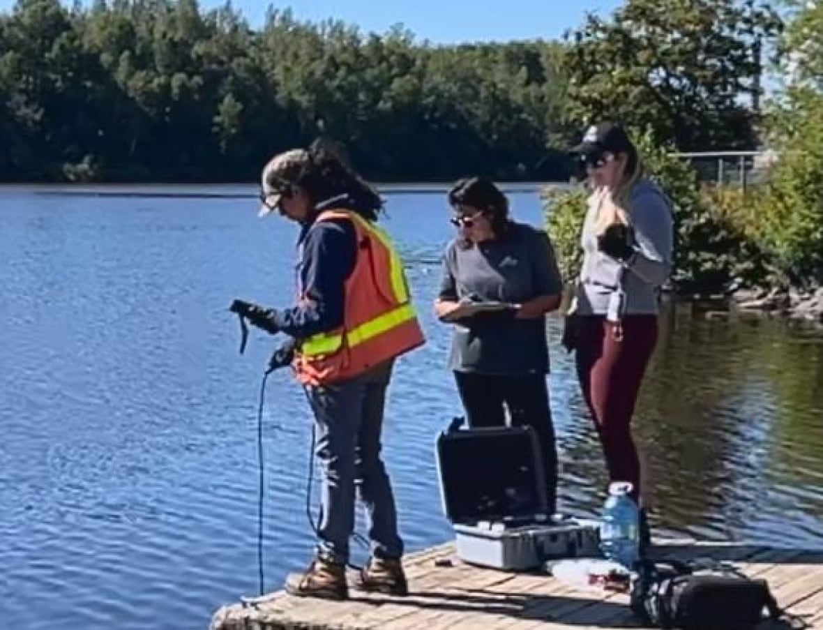 A woman in an orange safety vest holds a line into the water while standing on a wooden dock next to a small plastic brief case. Two women wearing gray stand alongside her.