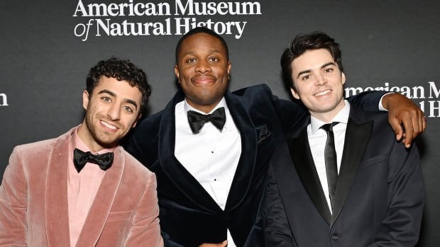 a man in a pink suit on the left and two men in black suits huddle together for a photo on a red carpet