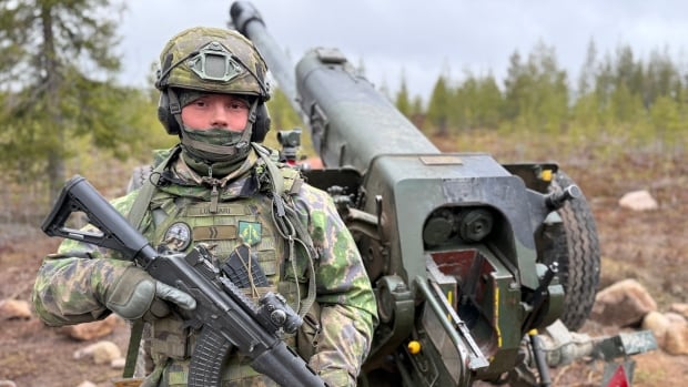 A young Finnish man holds a rifle next to a howitzer cannon. He's wearing green military tactical gear.