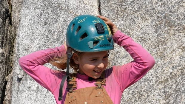 A girl with brown hair and wearing a pink shirt puts on a blue helmet. 