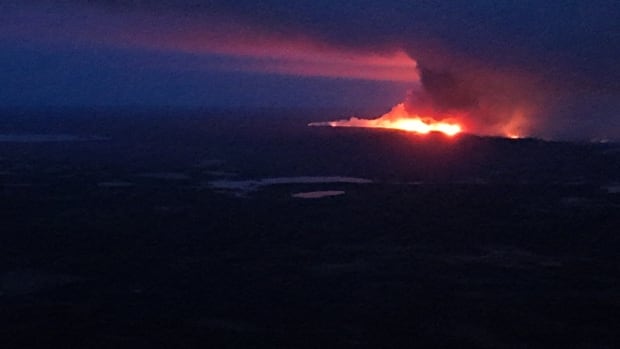 Aerial shot of a nearly pitch-black forest landscape with a raging fire illuminating a sky full of smoke in the distance. 