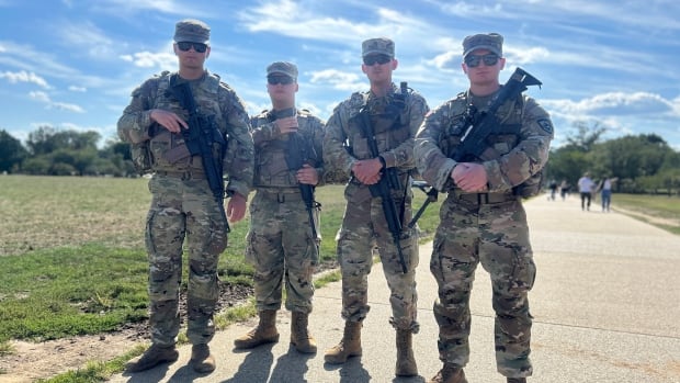 Four National Guards men in fatigues. They are all wearing hats and sunglasses carrying large rifles. Standing together. The sky is blue and there are a few clouds.