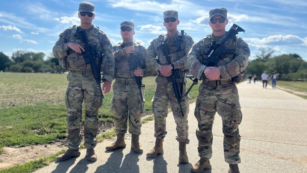 Four National Guards men in fatigues. They are all wearing hats and sunglasses carrying large rifles. Standing together. The sky is blue and there are a few clouds.