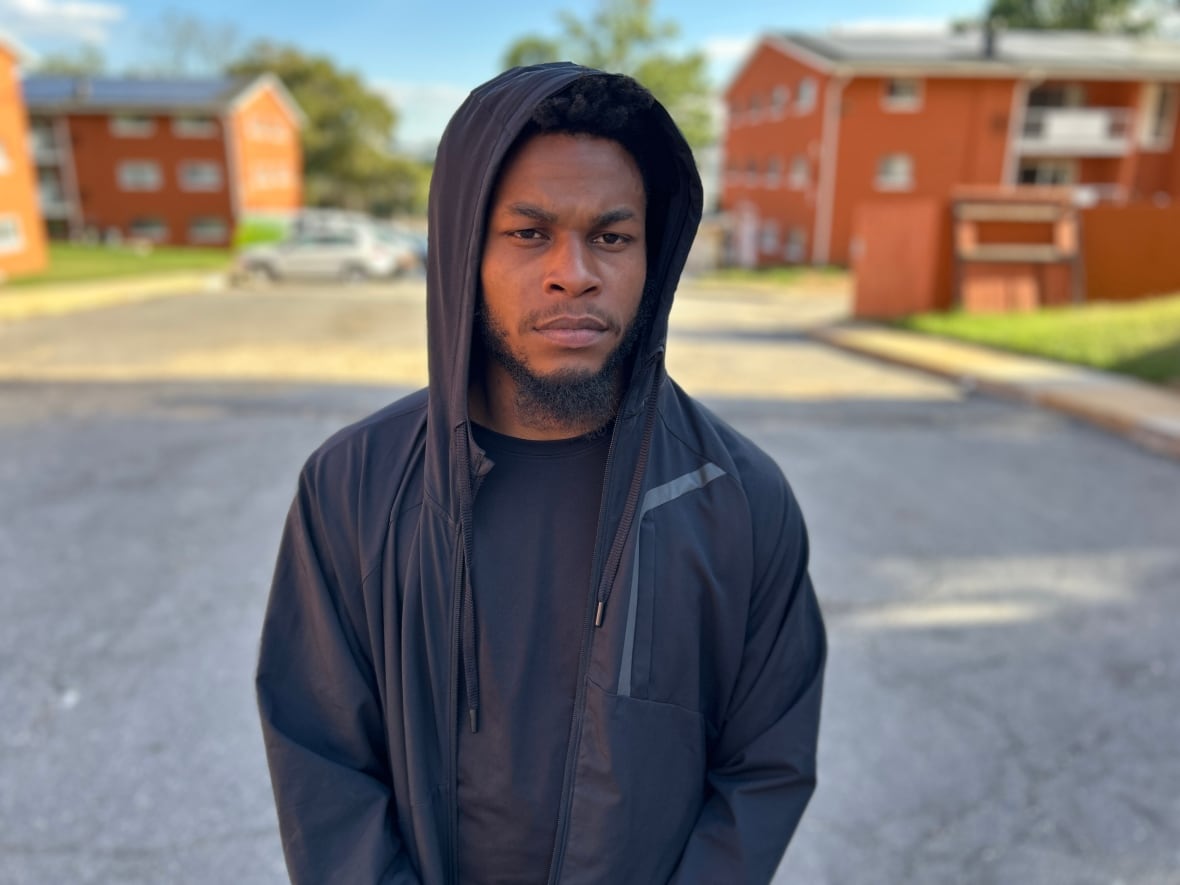 Brian Turner is a black man with short black hair and employed. He wears a black nylon jacket with a headscarf. He has a black t -shirt and stands in the midst of a car park. You can see three buildings of red brick behind it.