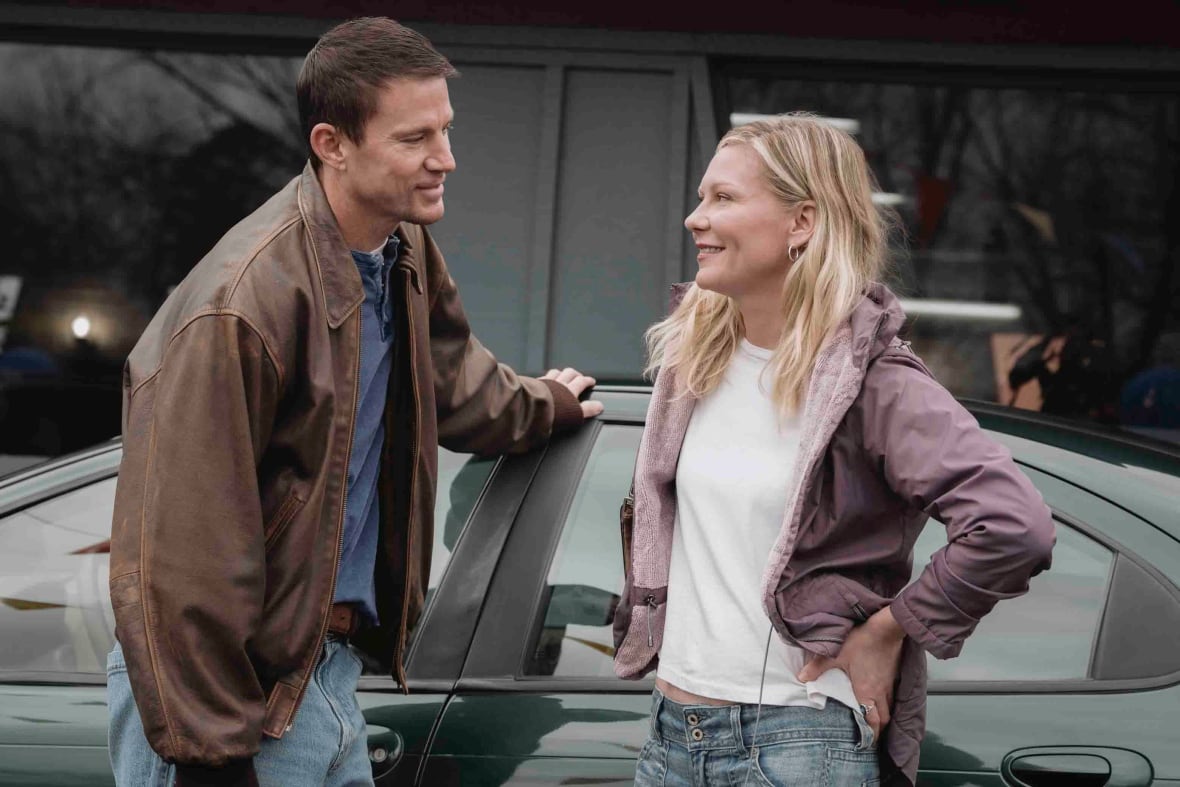 A smiling man and woman lean against a car while looking at one another.