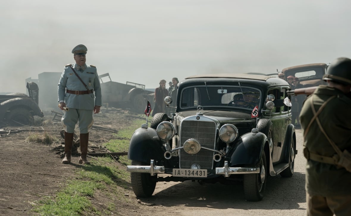 A man in a military uniform walks beside an old fashioned car. 