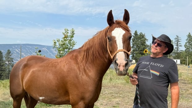 John Molund, a caucasian man in a hat, stands beside his brown horse Oakley 