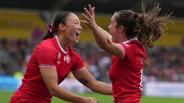 Two female rugby players representing Canada smile while moving in to wrap their arms around each other in celebration.
